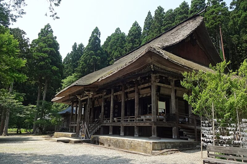 Temple traditionnel en bois au toit de chaume, niché au milieu de grands conifères.