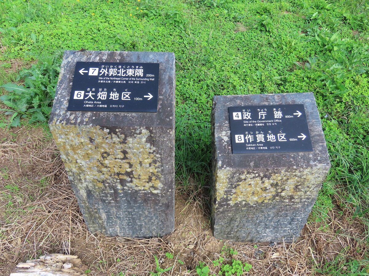 两根风化的石柱,上面有黑色的方向牌,标明距离(例如,到东北角 200 米,到大畑地区 100 米),周围环绕着绿色的植被和三叶草。.