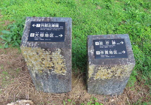 Two weathered stone posts with black directional plaques showing distances (eg, 200m to the Northeast Corner, 100m to Ohata Area) amid green vegetation and clover.