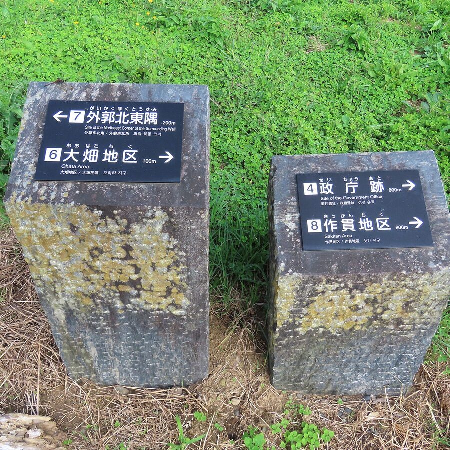 Two weathered stone posts with black directional plaques showing distances (e.g., 200m to the Northeast Corner, 100m to Ohata Area) amid green vegetation and clover.