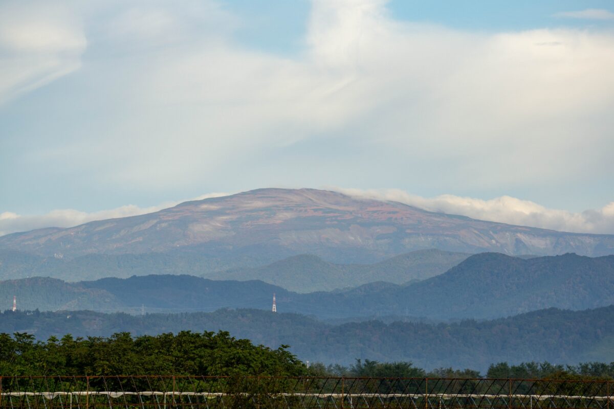Wide landscape of a mountain range under a blue sky with scattered clouds, a distant horizon and a barbed-wire fence in the foreground.