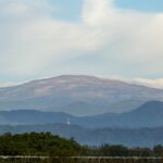 Amplio paisaje de una cadena montañosa bajo un cielo azul con nubes dispersas, un horizonte lejano y una cerca de alambre de púas en primer plano.