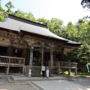 Traditional wooden Japanese temple with a curved roof surrounded by green trees; two visitors at the entrance.