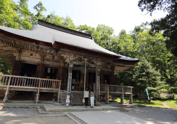 Traditional wooden Korean temple with a curved roof surrounded by green trees; two visitors at the entrance.