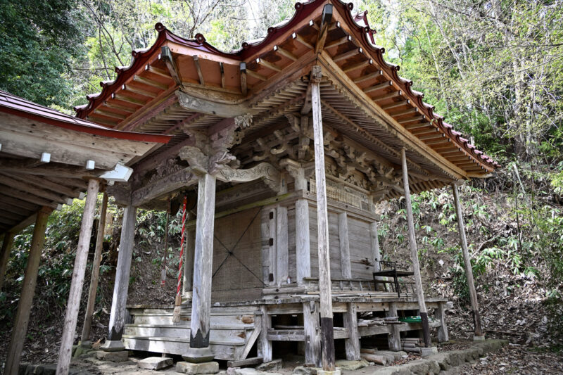 Traditional wooden shrine with decorated eaves and carved beams in a forest setting.