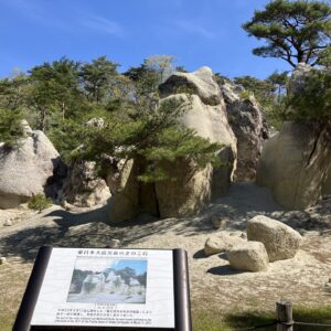 岩石嶙峋的地貌，巨大的岩石和松树在蓝天下若隐若现；前景中立着一块信息牌。.