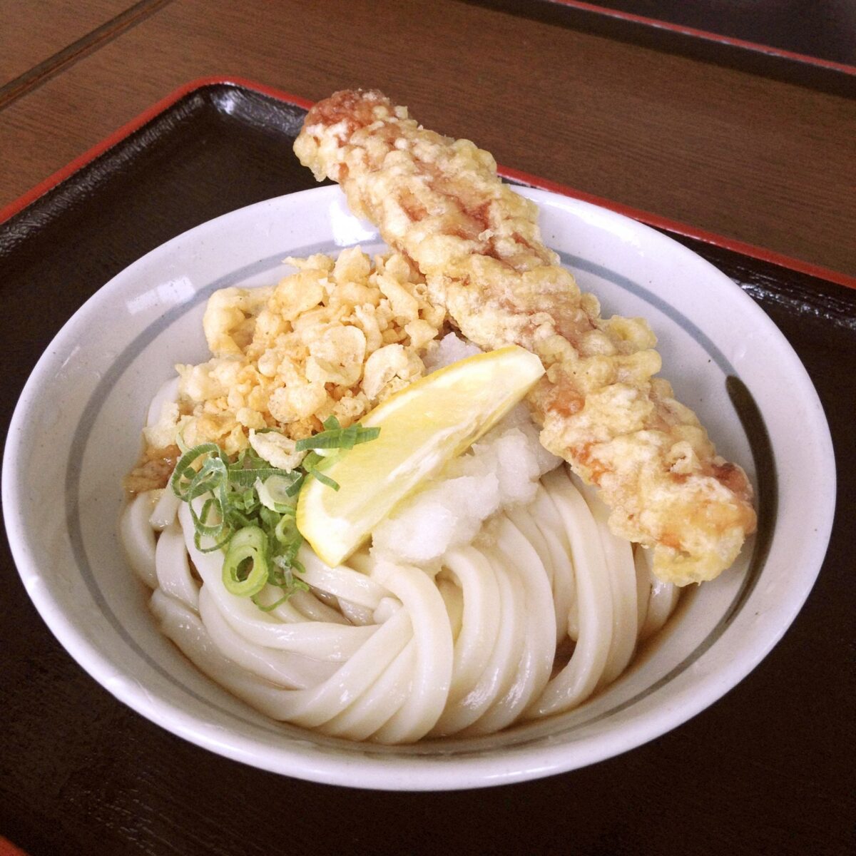 Un tazón de fideos udon gruesos con camarones tempura, una rodaja de limón y cebolletas picadas en caldo sobre una mesa de madera