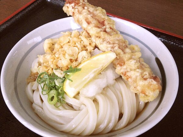 Bowl of thick udon noodles with tempura shrimp, a lemon wedge, and chopped green onions in broth on a wooden table