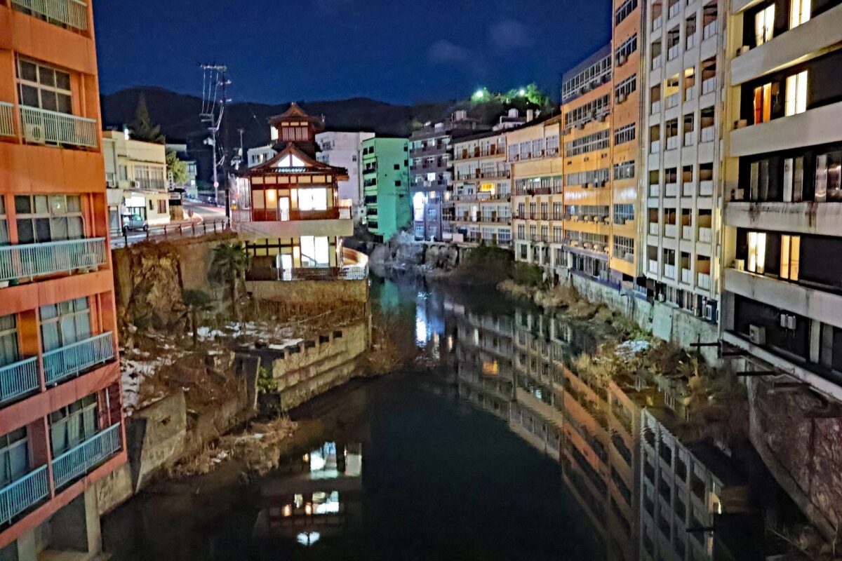 夜幕降临，城市运河中央矗立着一座灯火通明的传统建筑，倒映在水中，两侧是高耸的公寓楼。.