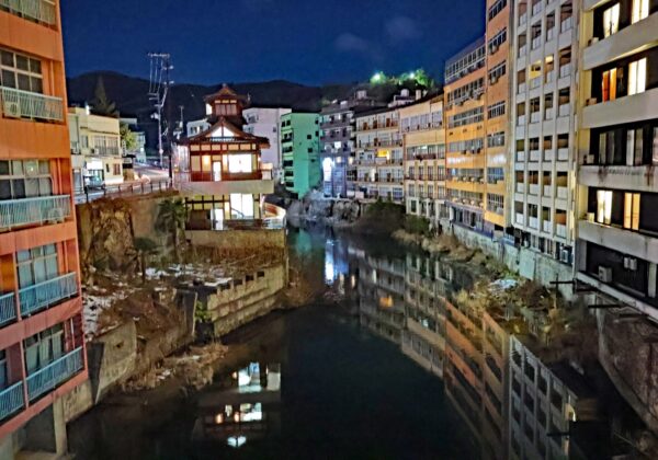 Canal nocturno de la ciudad con un edificio tradicional iluminado en el centro, reflejos en el agua, flanqueado por altos bloques de apartamentos.