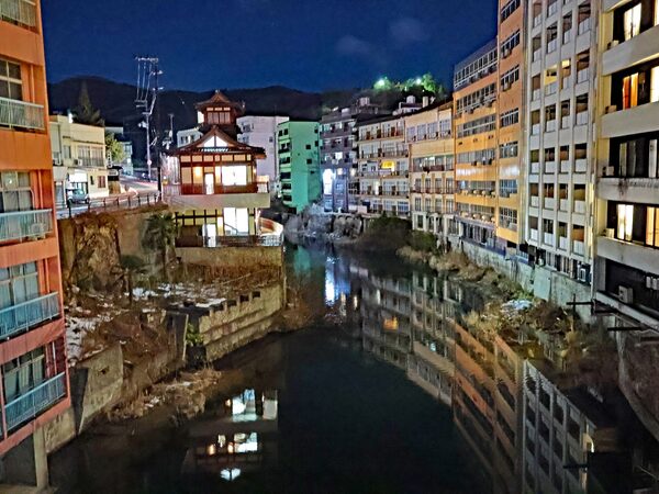 Nighttime city canal with a lit traditional building in the center, reflections on the water, flanked by tall apartment blocks.