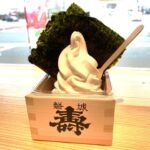 Vanilla soft-serve ice cream in a cup, topped with a large sheet of seaweed and a wooden spoon nearby on a wooden table.