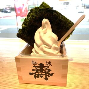 Vanilla soft-serve ice cream in a cup, topped with a large sheet of seaweed and a wooden spoon nearby on a wooden table.