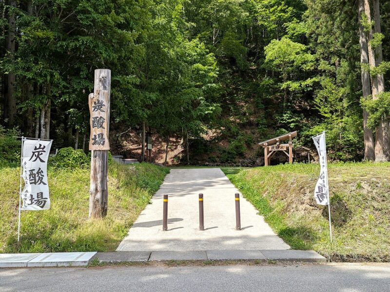 Trail entrance in a wooded area with a paved path, wooden posts, and white banners with Japanese characters on both sides.