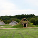 【縄文時代の青森③】豊かな森と海が育んだ縄文人の暮らし…人口約9000人の時代へ Thatched round huts on a green field with a dirt path and a tree-filled background.