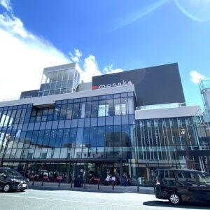 Modern glass-front shopping building with a gray upper block and a red heart logo, busy street in front with cars and pedestrians under a blue sky.