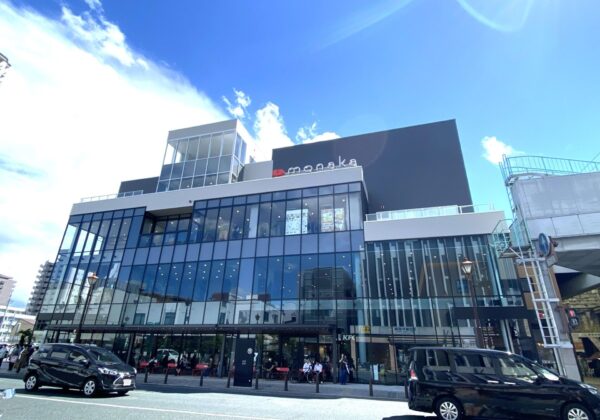 Modern glass-front shopping building with a gray upper block and a red heart logo, busy street in front with cars and pedestrians under a blue sky.
