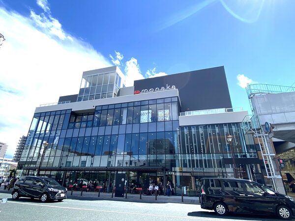 Modern glass-front shopping building with a gray upper block and a red heart logo, busy street in front with cars and pedestrians under a blue sky.