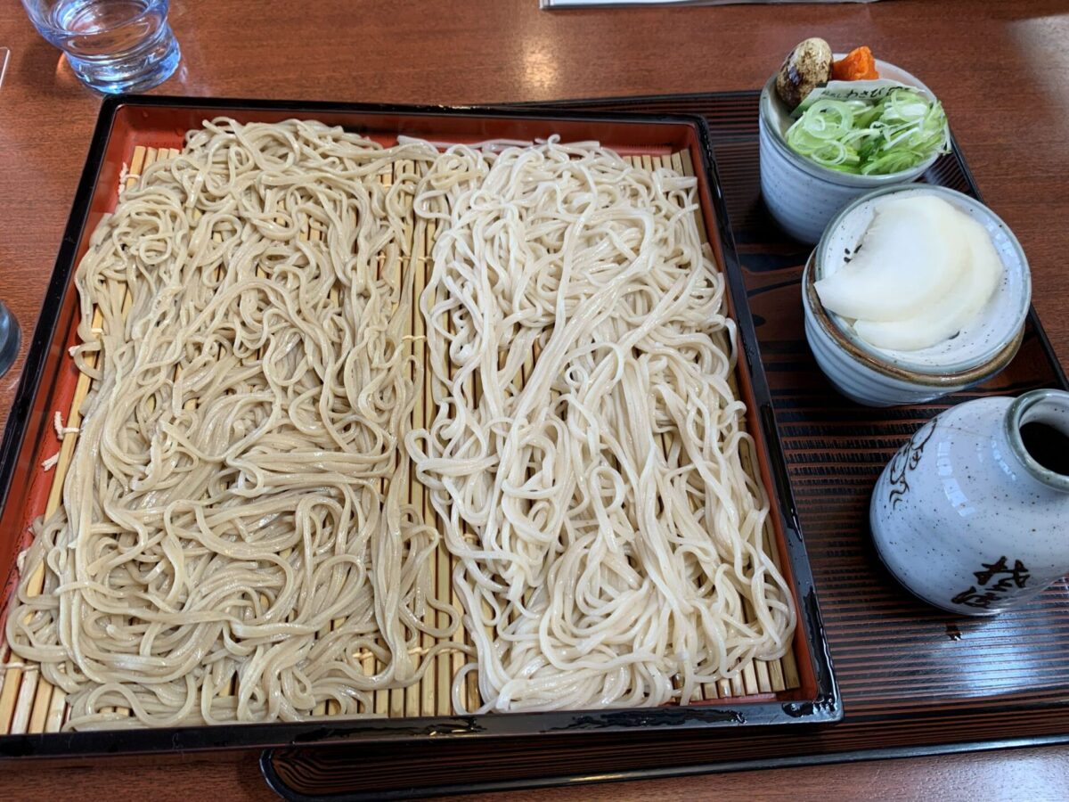 Cold soba noodles on a wooden mat in a lacquered tray, with green onion toppings and a side dish in bowls; glass of water nearby.