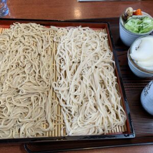 Cold soba noodles on a wooden mat in a lacquered tray, with green onion toppings and a side dish in bowls; glass of water nearby.