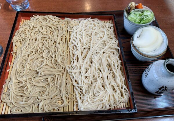 Cold soba noodles on a wooden mat in a lacquered tray, with green onion toppings and a side dish in bowls; glass of water nearby.