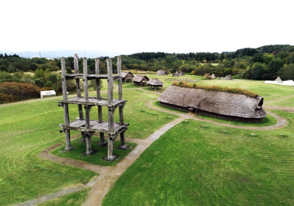 Vista aérea de un parque histórico de un pueblo con cabañas de techo de paja y una alta torre de madera en primer plano, con senderos curvos que atraviesan campos verdes