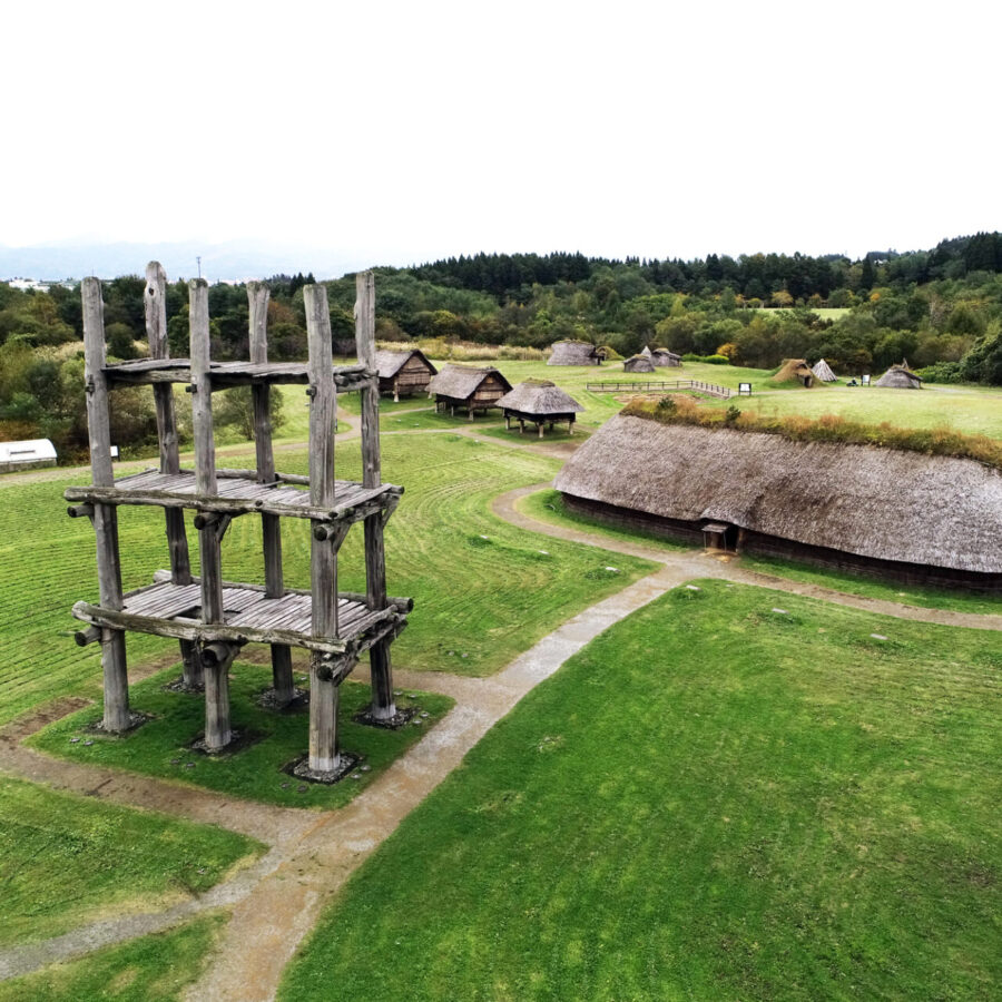 Aerial view of a historical village park with thatched huts and a tall wooden tower in the foreground, curving paths across green fields.]