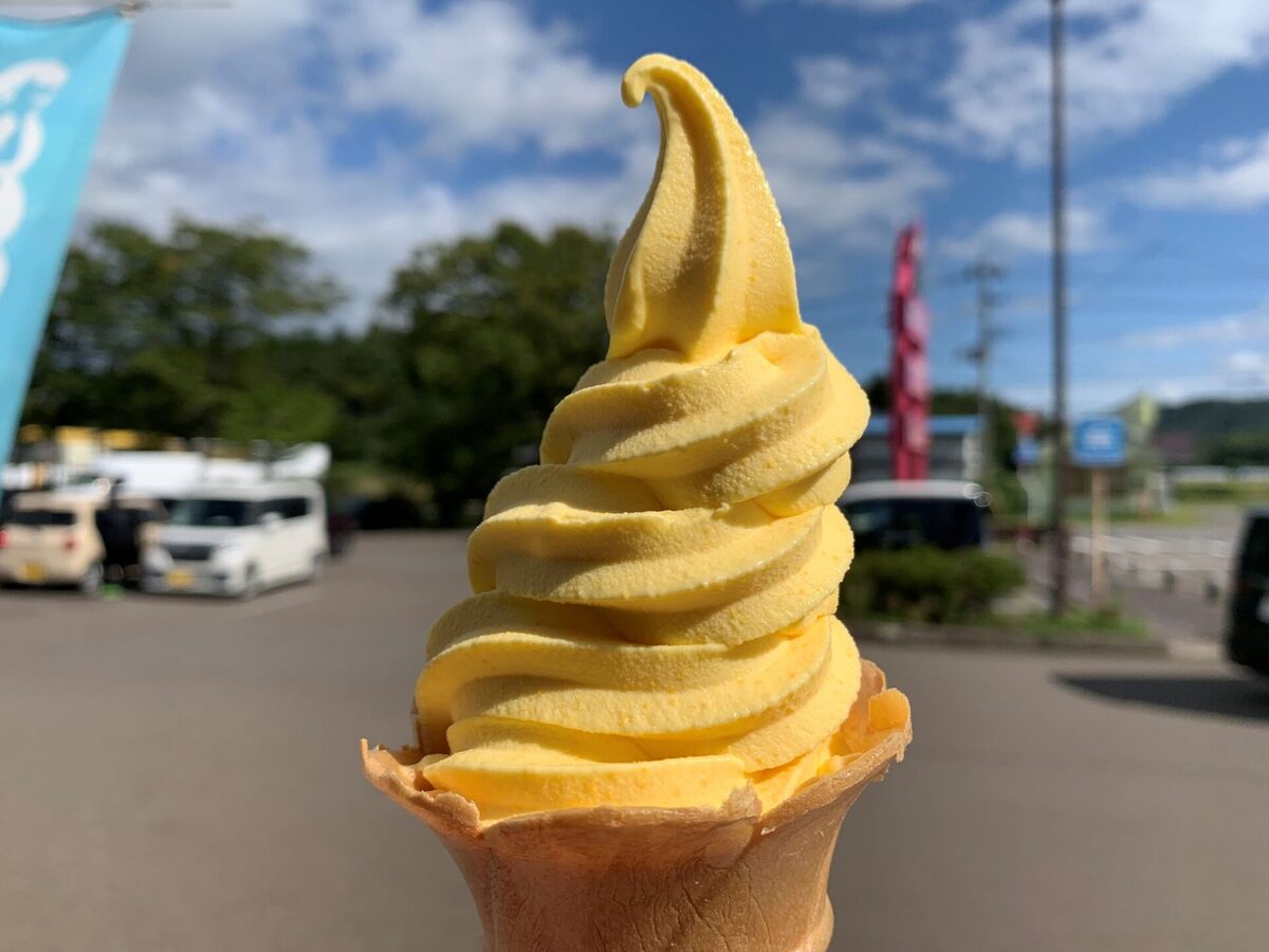 Yellow soft-serve ice cream cone held up outdoors on a sunny day, with a blurred parking lot background.