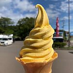 秋田県内陸北部の道の駅で味わう美味しいソフトクリーム!果実に野菜に卵風味、逆さにしても落ちないソフト!?【秋田県】 Yellow soft-serve ice cream cone held up outdoors on a sunny day, with a blurred parking lot background.