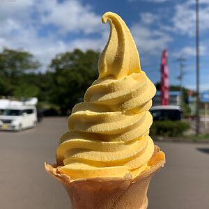 Yellow soft-serve ice cream cone held up outdoors on a sunny day, with a blurred parking lot background.