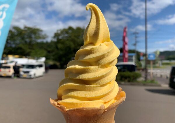 Yellow soft-serve ice cream cone held up outdoors on a sunny day, with a blurred parking lot background.