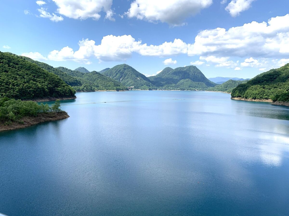 A calm blue lake bordered by forested green hills and distant mountains under a bright, partly cloudy sky.