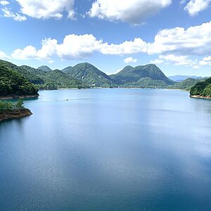 A calm blue lake bordered by forested green hills and distant mountains under a bright, partly cloudy sky.