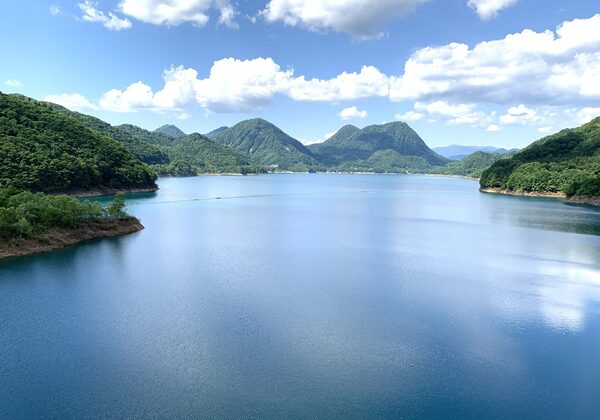 A calm blue lake bordered by forested green hills and distant mountains under a bright, partly cloudy sky.