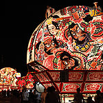 Festival nocturno con una gran carroza iluminada que presenta coloridos samuráis y figuras dramáticas contra el cielo oscuro.