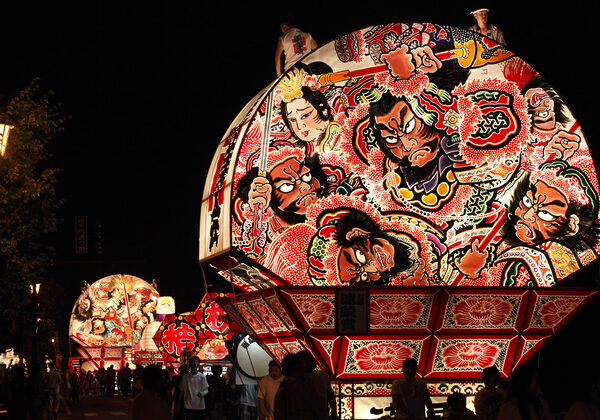 Night festival with a large illuminated float featuring colorful samurai and dramatic figures against the dark sky.