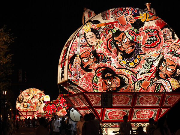 Night festival with a large illuminated float featuring colorful samurai and dramatic figures against the dark sky.
