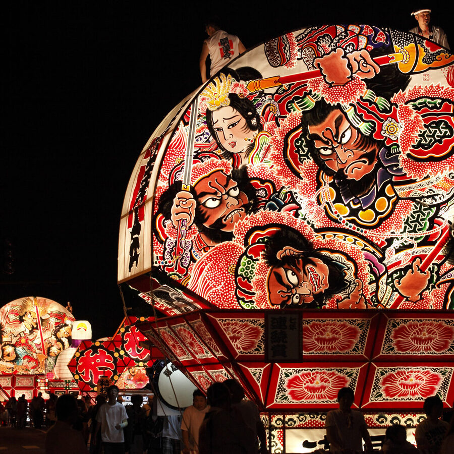 Night festival with a large illuminated float featuring colorful samurai and dramatic figures against the dark sky.