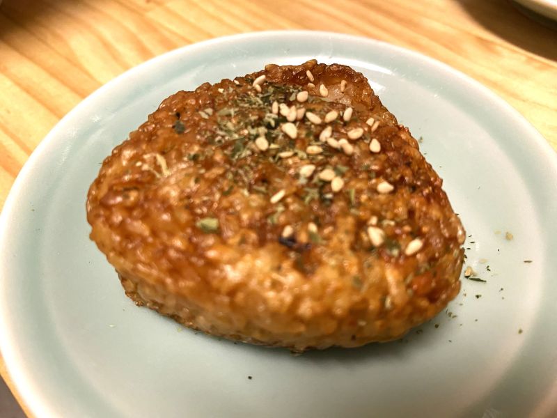 A slightly out-of-focus picture of a grilled rice ball topped with white sesame seeds and dried seaweed